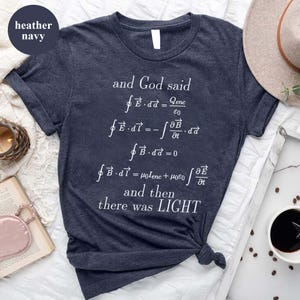 May include: Heather navy t-shirt with white text and mathematical equations. The text reads "and God said...and then there was LIGHT". The shirt is knotted at the bottom. A casual, everyday wear tee.
