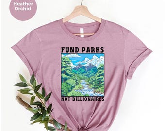 May include: Heather orchid-colored t-shirt with a graphic of a mountain landscape and the text "FUND PARKS NOT BILLIONAIRES". The design features a river, trees, and mountains under a blue sky. The shirt is hanging on a wooden hanger.
