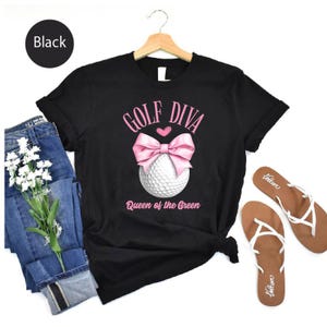 May include: Black t-shirt with a pink bow and golf ball graphic. The text reads "GOLF DIVA" above the bow and "Queen of the Green" below. The shirt is paired with blue jeans and brown sandals.