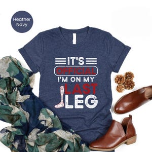 Puede incluir: Camiseta azul marino con el texto "IT'S OFFICIAL I'M ON MY LAST LEG" en blanco y rojo. El diseño incluye una ilustración de una pierna protésica. La imagen se presenta con zapatos marrones y una bufanda estampada.