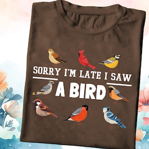 May include: Brown t-shirt with the text "Sorry I'm late I saw a bird" in white. The shirt features a variety of colorful birds, including a cardinal, a blue jay, and a sparrow.