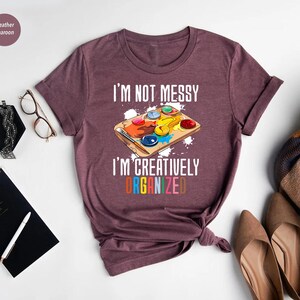 May include: Heather maroon t-shirt with the text "I'M NOT MESSY I'M CREATIVELY ORGANIZED" and a colorful paint palette graphic. The shirt has rolled-up sleeves and a crew neck. The text and graphic are in white and various colors.
