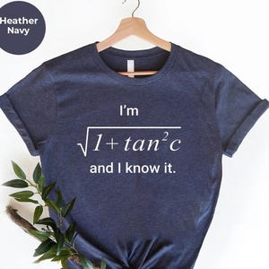 Puede incluir: Camiseta azul marino con el texto "I'm √(1+tan²c) and I know it." en blanco. La camiseta está colgada de una percha de madera. Un círculo azul oscuro con las palabras "Heather Navy" está en la esquina superior izquierda.