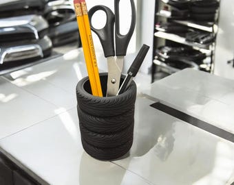 May include: A black tire-shaped pencil holder with four stacked tires. The holder contains two yellow pencils, a black pen, and a pair of black scissors.
