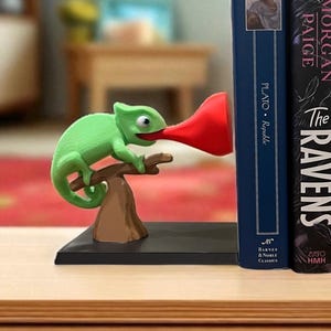 May include: A whimsical green chameleon bookend with a red horn-shaped tongue, perched on a brown branch. The bookend is placed next to two books, one blue and one black, on a wooden surface. The bookend adds a playful touch to any bookshelf.