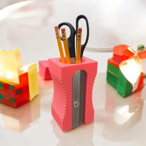 May include: A pink plastic pencil sharpener with a built-in pencil holder. Three yellow pencils and a pair of black scissors are in the holder.