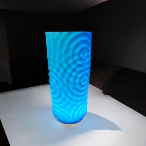 May include: A tall, cylindrical blue lamp with a textured surface featuring concentric circle patterns. The lamp emits a bright, cool blue light, creating a soft glow. The lamp is on a white surface.