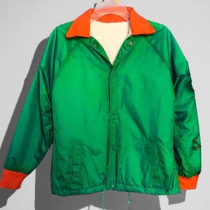 May include: A green jacket with an orange collar and cuffs. The jacket has a snap closure and two front pockets. The cuffs have a decorative design. The jacket is hanging on a hanger against a gray background.