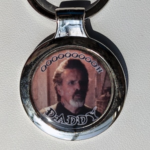 May include: Silver keychain with a round, silver-colored charm featuring a photo of a man with the word "Daddy" printed below the photo. The charm has a silver ring around the edge with the word "Ooooooooh" printed around the ring.