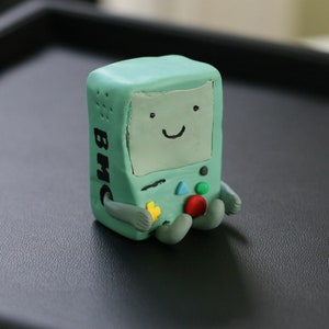 Puede incluir: Una figurita artesanal verde y azul de un personaje de dibujos animados que se asemeja a una consola de juegos. El personaje tiene una cara sonriente y un botón en el pecho que dice "BMO".