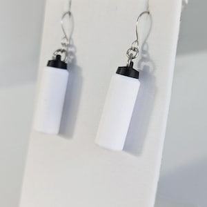 May include: A pair of white and black earrings shaped like miniature water bottles. The earrings have silver-colored hooks and are displayed on a white stand. The water bottle earrings are a unique accessory.