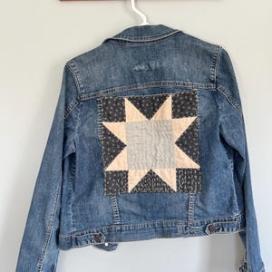 May include: A blue denim jacket with a star-shaped quilt patch on the back. The patch features a light beige center, surrounded by gray and black patterned fabric. The jacket is hanging on a white hanger.