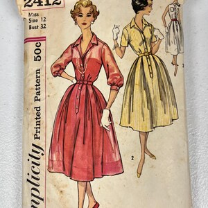 May include: A vintage Simplicity sewing pattern envelope, number 2412, for a Miss size 12, bust 32 dress. The illustration features two variations of a collared, button-front dress with a full skirt, one in solid red and one in yellow stripes.
