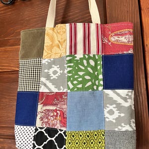 May include: A rectangular tote bag constructed from a patchwork of various fabric squares. The design features diverse patterns including red and white stripes, black and white gingham, polka dots, floral motifs, and solid colors in blue, green, and gray.