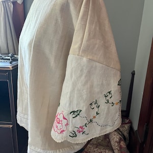 May include: A cream-colored linen blouse featuring wide, flared sleeves with intricate floral embroidery in pink, green, and yellow. The garment includes a scalloped lace trim along the sleeve edges and a delicate hemline detail at the bottom.