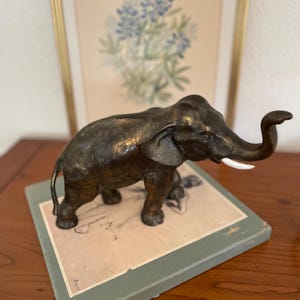 May include: A bronze-toned elephant figurine standing on a square, light green base. The elephant has a textured, dark metallic finish, large ears, and a single visible white tusk curving upward. The trunk is raised, and the tail hangs down.