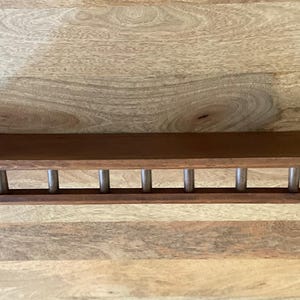 May include: A rectangular, dark brown wooden shelf with a series of evenly spaced, silver-colored metal rods beneath. The shelf is mounted on a light brown wooden surface.