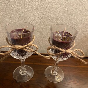 May include: Two wine glass candles with a burgundy colored wax and gold glitter. The candles are tied with twine and a bow.