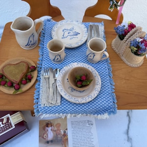 May include: A miniature dining set featuring a blue and white checkered runner, two ceramic mugs, a pitcher, and two plates with blue bird motifs. Includes a heart-shaped pastry on a plate, a floral centerpiece, metal cutlery, and a small wooden bowl.