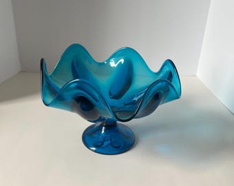 Könnte beinhalten: Eine leuchtend türkisfarbene Glasschale mit gerüschtem Rand, die auf einem Sockel steht. Das wellenförmige Design und die satte Farbe der Schale erzeugen einen auffälligen visuellen Reiz. Dieses dekorative Stück ist ideal für die Präsentation von Obst oder als Mittelstück.
