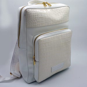 May include: White leather backpack with a crocodile embossed design. The backpack has gold zippers and a small pocket on the front.