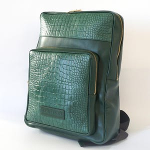 May include: A dark green leather backpack with a crocodile embossed pattern. The backpack has a large front pocket with a gold zipper and the brand name "MAURUMON" embossed on the front.