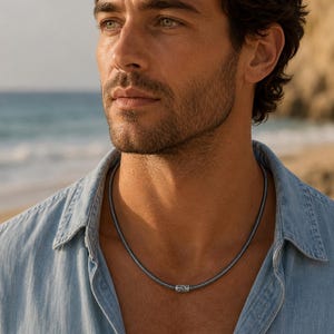 May include: A dark grey metallic snake chain necklace featuring a single cylindrical bead with an intricate filigree pattern. The necklace rests around the neck, showcasing a textured, flexible link design that provides a sleek and uniform appearance.