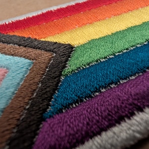 May include: A rectangular embroidered patch featuring the Progress Pride flag design. The patch displays horizontal stripes in red, orange, yellow, green, blue, and purple, with a chevron on the left side containing light blue, pink, white, brown, and black.