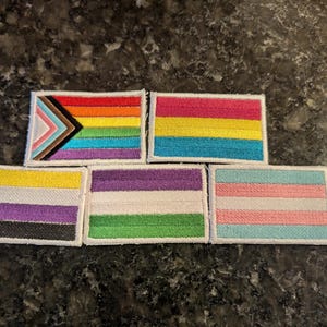 May include: Five rectangular embroidered patches featuring various pride flags. The top row shows the Progress Pride flag and the pansexual flag. The bottom row displays the non-binary flag, the genderqueer flag, and the transgender flag.