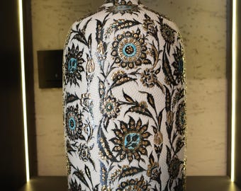 Può includere: Un vaso in ceramica bianca con un bordo dorato e intricati disegni floreali neri e dorati. Il vaso ha un corpo rotondo e un collo stretto.
