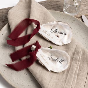 Könnte beinhalten: Zwei Austernschalen als Platzkarten, jeweils mit einem handgeschriebenen Namen, Thomas und Isabell, in schwarzer Schrift. Ein kleines weißes Schild mit der Aufschrift Thank you ist mit einem dunkelroten, ausgefransten Band befestigt.