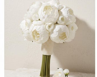 Puede incluir: Un ramo de peonías blancas con pétalos en capas y tallos verdes, con un mango envuelto en una textura de cuentas blancas. Una sola flor de peonía blanca a juego con un tallo verde se coloca por separado frente al ramo principal.