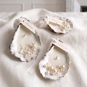 May include: Three oyster shell candles filled with white wax and a single wick. Each shell is decorated with various sizes of white pearls and features a small, cream-coloured paper tag tied with a white ribbon that says Thank you in script.