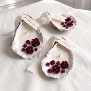 Può includere: Tre candele in guscio di ostrica riempite di cera bianca, decorate con piccoli accenti di cera a forma di rosa rosso scuro. Ogni guscio presenta uno stoppino centrale e un cartellino di carta color crema legato con un nastro bianco, con la scritta Thank you.