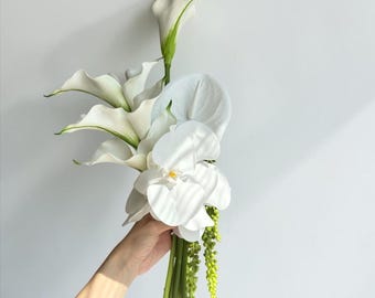 Puede incluir: Un ramo de flores que presenta lirios de cala blancos, un anturio blanco y una gran orquídea blanca con un centro amarillo. El arreglo incluye tallos verdes y pequeños racimos de flores verdes colgantes, todos reunidos en un solo manojo.
