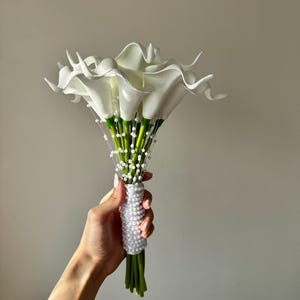 May include: A bouquet of white calla lilies with green stems, accented by thin strands of small white beads. The handle of the bouquet is wrapped in a dense layer of white faux pearls, creating a textured, cylindrical grip for the floral arrangement.