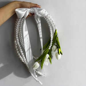 Peut inclure: Un serre-tête blanc orné de fausses perles et d'un nœud en satin. Le serre-tête est orné de fleurs de tulipes blanches et de feuilles vertes. Les perles sont disposées en plusieurs brins, créant un effet superposé. Le nœud et les rubans ajoutent une touche d'élégance.