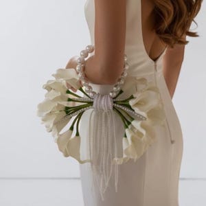 May include: A floor-length white sleeveless dress with a low back, paired with a circular bouquet of white calla lilies. The bouquet features a handle made of large white pearls and is accented with long, dangling strands of delicate white beads.