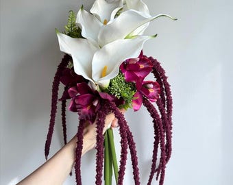 Puede incluir: Un ramo de flores con calas blancas de centro amarillo, orquídeas color magenta intenso y largas hebras colgantes de amaranto púrpura oscuro. El arreglo incluye acentos de follaje verde y tallos verdes agrupados en la base.