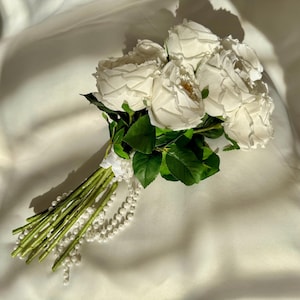 Könnte beinhalten: Ein Strauß aus weißen Rosen mit grünen Blättern und Stielen, dessen Griff mit weißem Stoff umwickelt ist und mit einer langen, herabhängenden Schnur aus weißen Kunstperlen verziert ist, die von der Basis der Blumenstiele herabfällt.