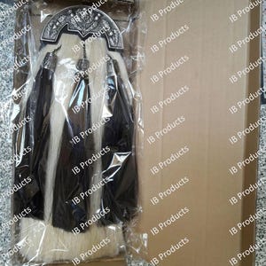 Puede incluir: Un sporran tradicional, un accesorio de vestimenta escocesa de las Highlands, se muestra en una bolsa de plástico transparente dentro de una caja de cartón. El sporran presenta un cantle plateado, borlas negras y un cuerpo de piel blanco y negro.
