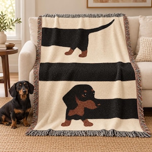 May include: A woven throw blanket featuring a cream background with wide horizontal black stripes. The design displays a black and tan dachshund with its body extending across the stripes, finished with a multicolored fringed edge.