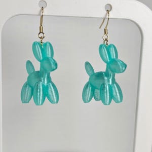 Peut inclure: Une paire de boucles d'oreilles turquoise imprimées en 3D en forme de chiens ballons. Les boucles d'oreilles ont des crochets d'oreilles dorés et sont présentées sur un fond blanc. Le design du chien ballon comprend une tête, un corps et quatre pattes.