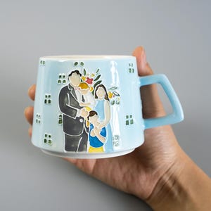 Puede incluir: Una taza de cerámica azul claro con una ilustración en relieve y colorida de una familia de cuatro. La taza tiene un asa geométrica y pequeños cuadrados verdes dispersos por la superficie. El retrato familiar incluye figuras con ropa oscura y clara.