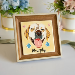 May include: A square ceramic tile featuring a raised, colourful illustration of a yellow Labrador retriever's face with its tongue out, surrounded by small pink hearts and blue flowers, with the name Murphy written below, set in a wooden frame.