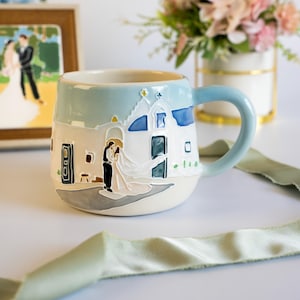 May include: A ceramic mug featuring a raised, hand-painted scene of a couple in wedding attire standing before a white building with blue domes. The mug has a light blue rim and handle, with a cream-coloured base and a grey ground illustration.