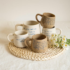May include: Four ceramic mugs in cream and tan colours, each featuring handwritten blue text. The mugs have rounded handles and irregular shapes. One mug displays the text "the story isn't over there is ending" and another says "I love you times infinity".