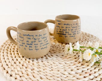 May include: Two beige ceramic mugs with handwritten blue text. The mugs have a rounded shape with a handle. The text on the mugs includes phrases like "Here's something to dry your day" and "I love you always". The mugs are on a woven placemat.