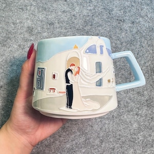 以下が含まれることがあります： 水色の持ち手と縁が付いたセラミックマグカップで、結婚式のシーンが描かれています。デザインには、青いアクセントのある白い建物の前で抱き合うカップルが含まれています。マグカップは手で持たれています。