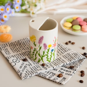 May include: A white ceramic mug decorated with a floral pattern of yellow, purple, and pink flowers. The mug rests on a newspaper-print napkin, with coffee beans scattered around. Macarons are in the background.